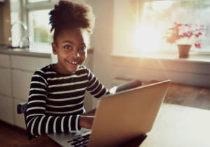 Smiling friendly young black african girl with a cute top knot afro hairstyle sitting at a laptop computer at the dining table grinning at the camera