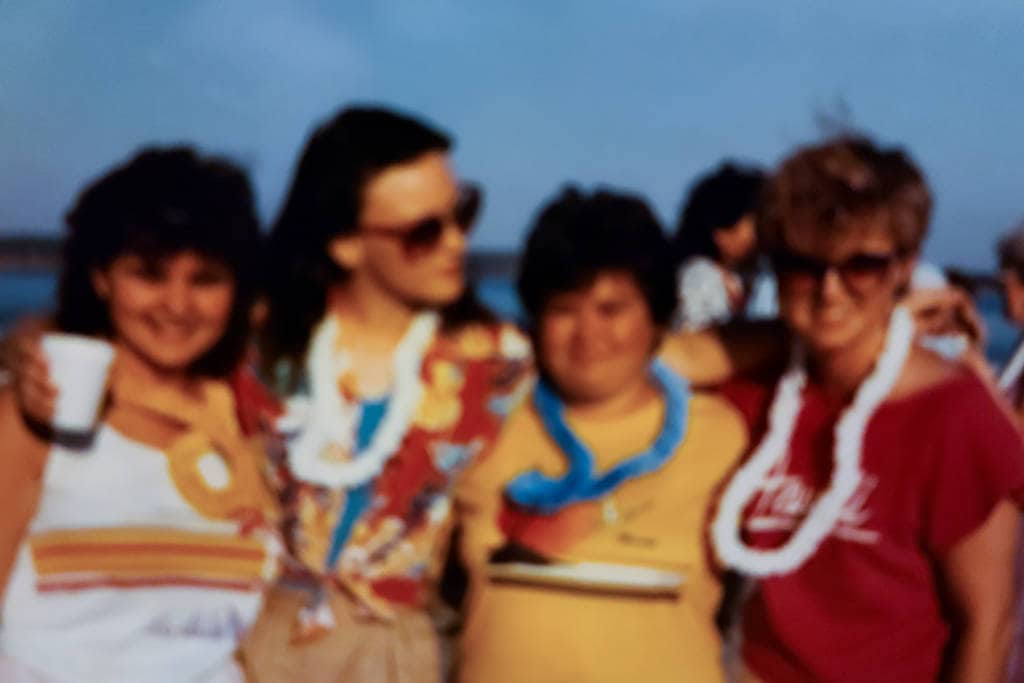 Group of four women in late 70s/early 80s clothes with Hawaiian lays around their necks, arms around each other smiling