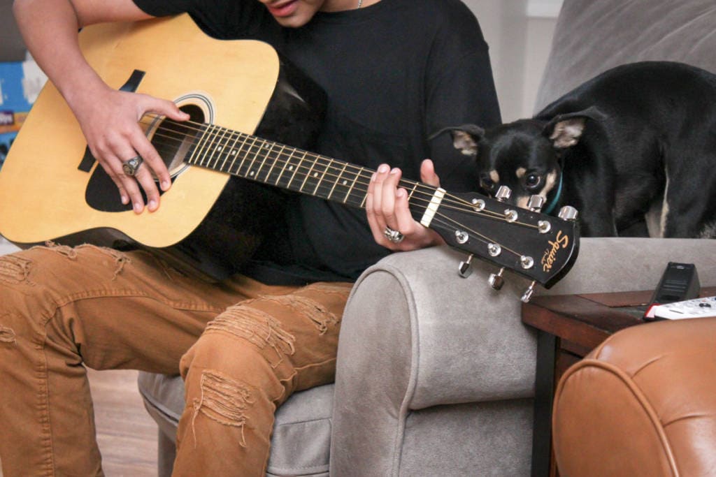 teenage boy playing guitar while his small dog watches on