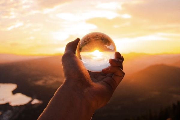 silhouetted hand holding up clear glass ball to sunset, light shining through