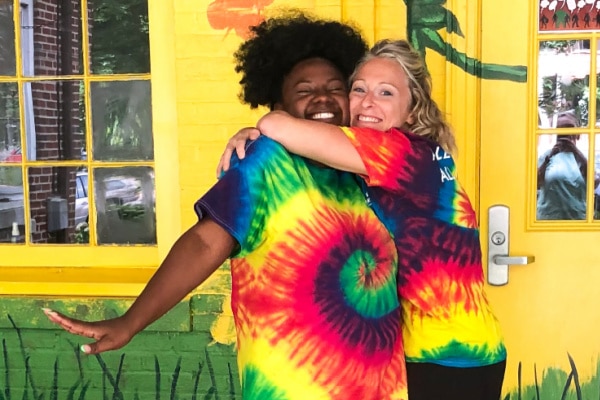 a black woman and a white woman, both wearing tie dyed rainbow t-shirts hug outside of a colorfully painted building
